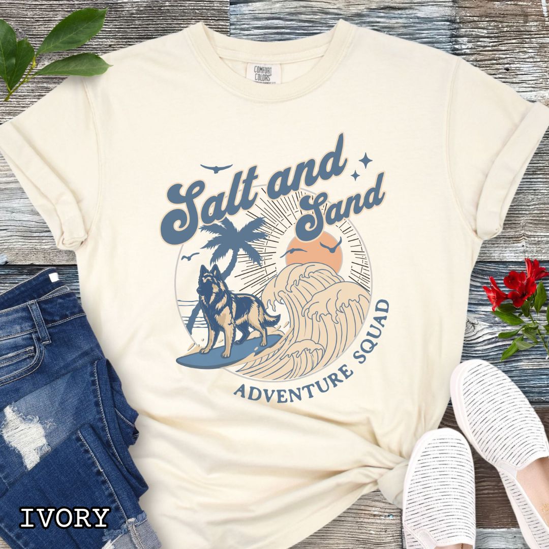 Beige Comfort Colors t-shirt for Belgian Tervuren lovers with beach-themed graphic of a surfing Belgian Tervuren, waves and sunset, and the text 'Salt and Sand Adventure Squad' in retro lettering, shown on a wooden surface with accessories.