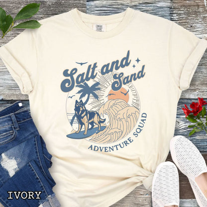 Beige Comfort Colors t-shirt for Belgian Tervuren lovers with beach-themed graphic of a surfing Belgian Tervuren, waves and sunset, and the text 'Salt and Sand Adventure Squad' in retro lettering, shown on a wooden surface with accessories.