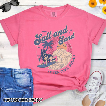 Bright pink Comfort Colors t-shirt for Belgian Tervuren lovers with beach-themed graphic of a surfing Belgian Tervuren, waves and sunset, and the text 'Salt and Sand Adventure Squad' in retro lettering, shown on a wooden surface with accessories.