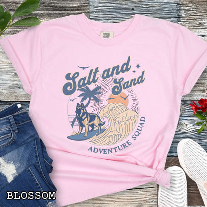 Pink Comfort Colors t-shirt for Belgian Tervuren lovers with beach-themed graphic of a surfing Belgian Tervuren, waves and sunset, and the text 'Salt and Sand Adventure Squad' in retro lettering, shown on a wooden surface with accessories.