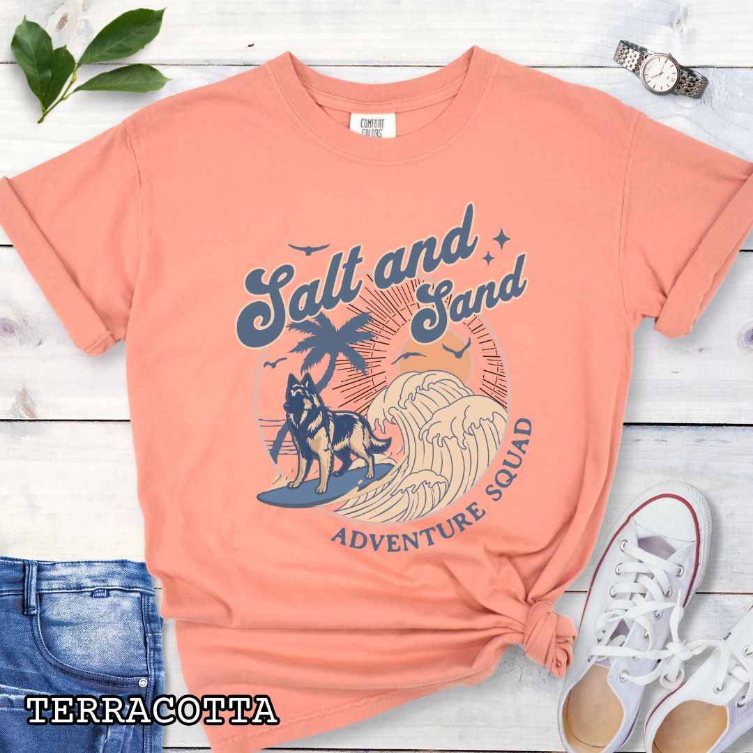 Orange Comfort Colors t-shirt for Belgian Tervuren lovers with beach-themed graphic of a surfing Belgian Tervuren, waves and sunset, and the text 'Salt and Sand Adventure Squad' in retro lettering, shown on a wooden surface with accessories.