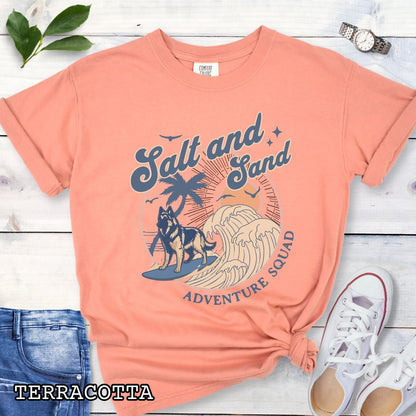 Orange Comfort Colors t-shirt for Belgian Tervuren lovers with beach-themed graphic of a surfing Belgian Tervuren, waves and sunset, and the text 'Salt and Sand Adventure Squad' in retro lettering, shown on a wooden surface with accessories.