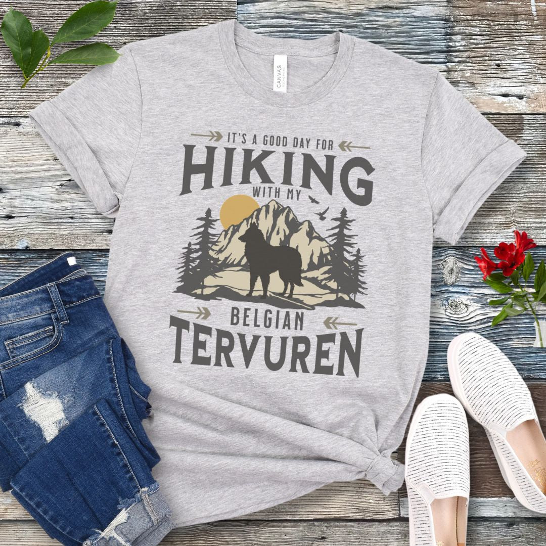 Grey Belgian Tervuren t-shirt with vintage-style mountain wilderness graphic, Tervuren dog silhouette and "Hiking With My Belgian Tervuren' text on a wooden surface with jeans and shoes.