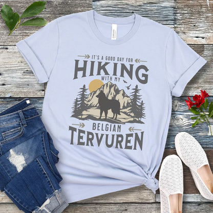 Light blue Belgian Tervuren t-shirt with vintage-style mountain wilderness graphic, Tervuren dog silhouette and "Hiking With My Belgian Tervuren' text on a wooden surface with jeans and shoes.