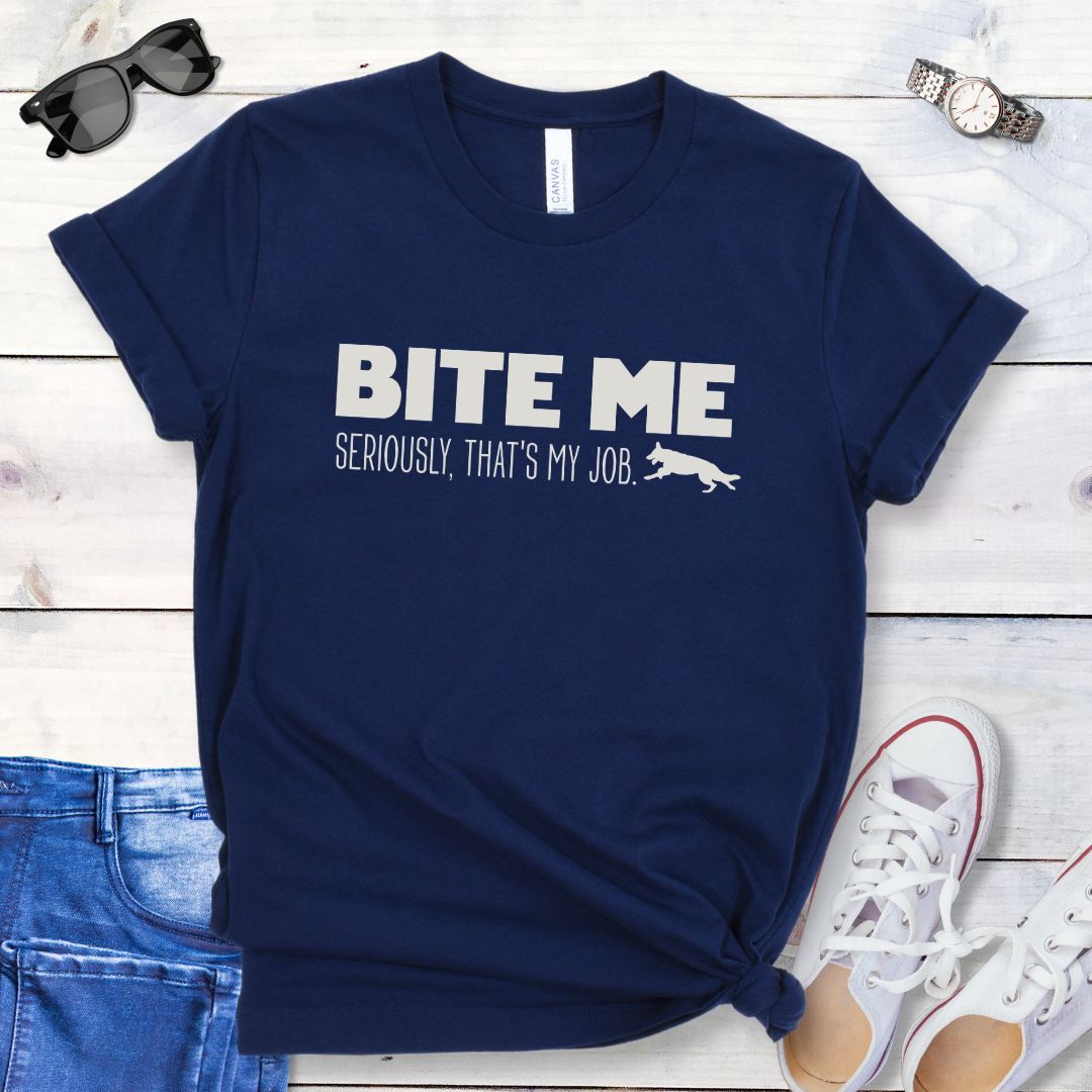 Navy blue K9 helper t-shirt with 'BITE ME Seriously, That's My Job' text and running dog graphic, presented on a wooden surface with jeans and shoes.