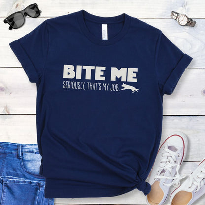 Navy blue K9 helper t-shirt with 'BITE ME Seriously, That's My Job' text and running dog graphic, presented on a wooden surface with jeans and shoes.