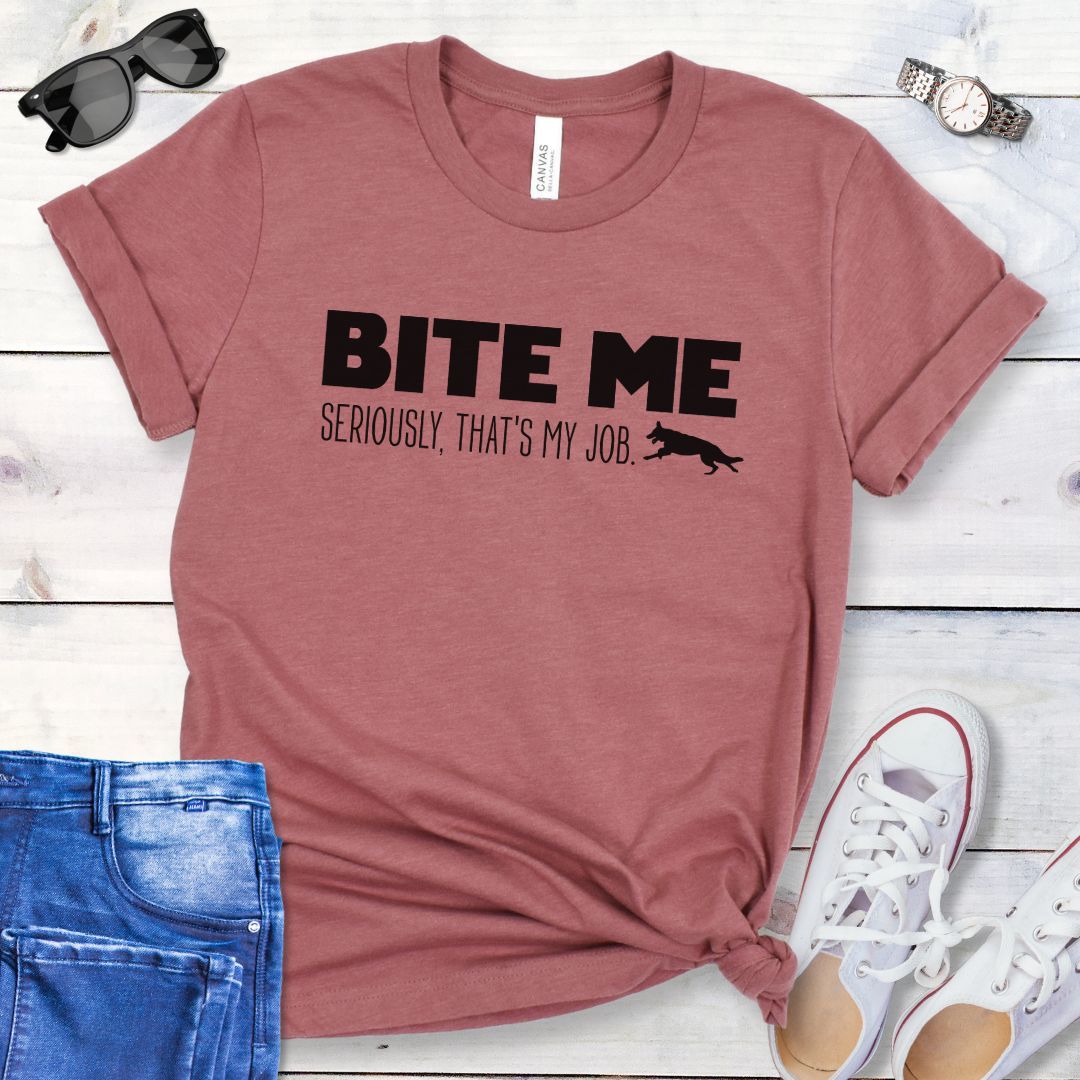 Mauve K9 helper t-shirt with 'BITE ME Seriously, That's My Job' text and running dog graphic, presented on a wooden surface with jeans and shoes.