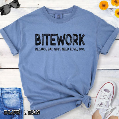 Blue Comfort Colors t-shirt for protection dog sports enthusiasts with humorous text 'BITEWORK BECAUSE BAD GUYS NEED LOVE, TOO.', shown on a wooden surface with jeans and sneakers.