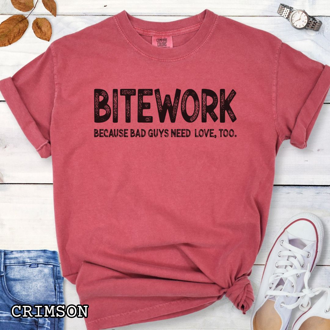 Red Comfort Colors t-shirt for protection dog sports enthusiasts with humorous text 'BITEWORK BECAUSE BAD GUYS NEED LOVE, TOO.', shown on a wooden surface with jeans and sneakers.