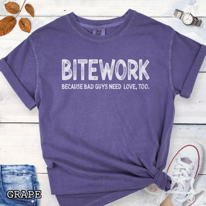 Purple Comfort Colors t-shirt for protection dog sports enthusiasts with humorous text 'BITEWORK BECAUSE BAD GUYS NEED LOVE, TOO.', shown on a wooden surface with jeans and sneakers.