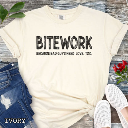 Beige Comfort Colors t-shirt for protection dog sports enthusiasts with humorous text 'BITEWORK BECAUSE BAD GUYS NEED LOVE, TOO.', shown on a wooden surface with jeans and sneakers.