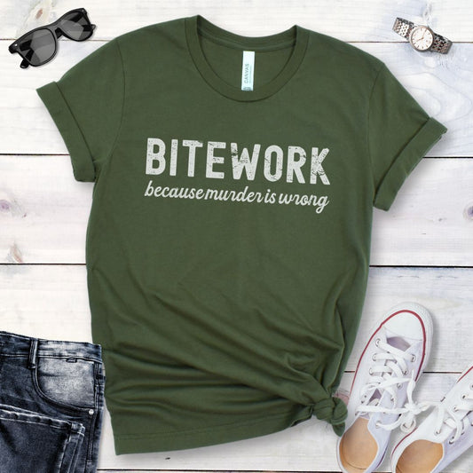 Military green Schutzhund/IGP dog sport t-shirt with humorous text 'BITEWORK because murder is wrong' in bold grunge lettering, shown on a wooden surface with jeans and sneakers. 