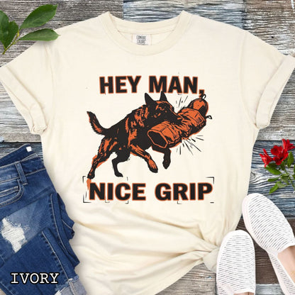 Ivory Comfort Colors t-shirt with graphic design of a Schutzhund dog biting a sleeve and the humorous text 'Hey Man nice grip', shown on a wooden surface with accessories.