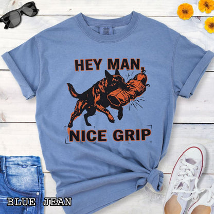 Blue Comfort Colors t-shirt with graphic design of a Schutzhund dog biting a sleeve and the humorous text 'Hey Man nice grip', shown on a wooden surface with accessories.