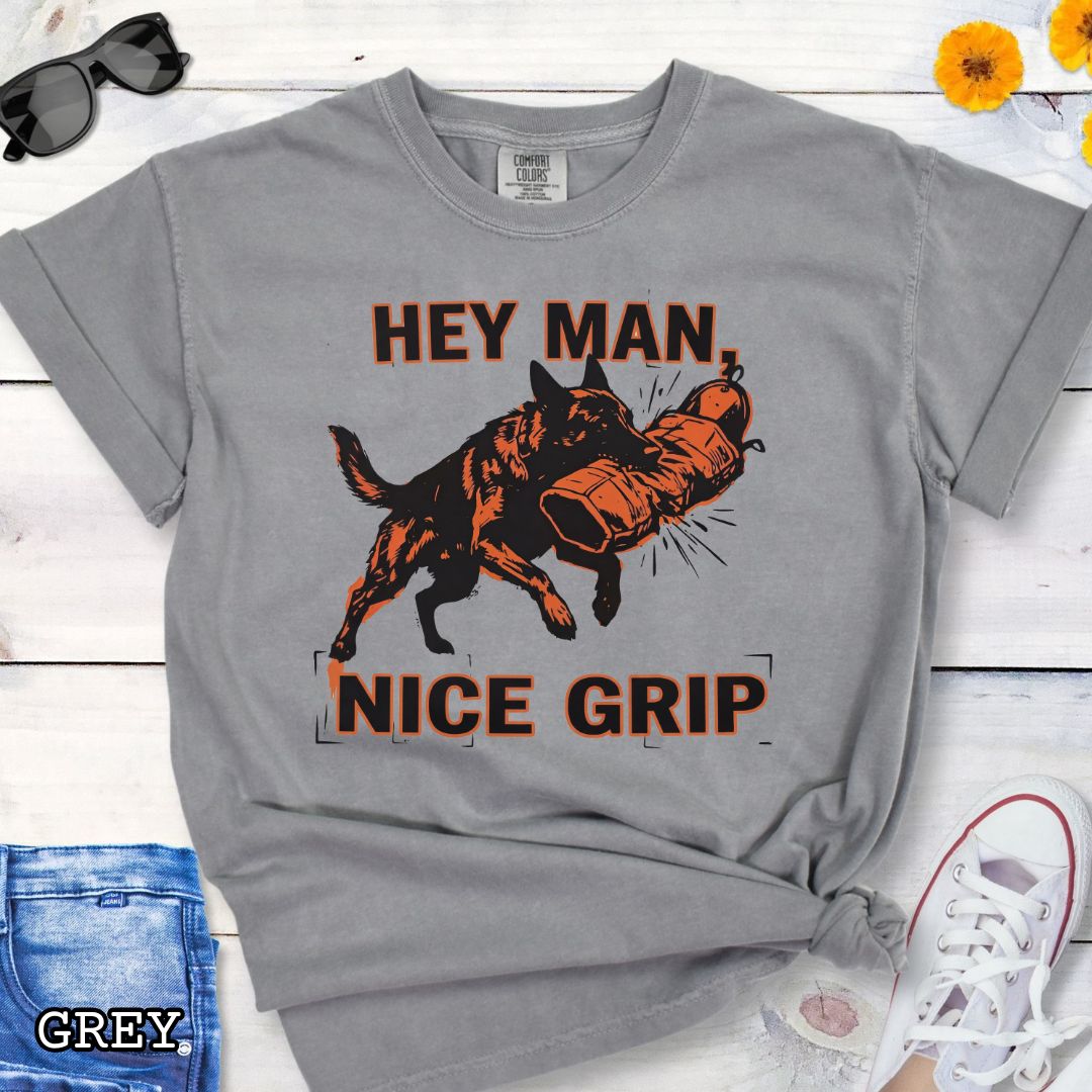Grey Comfort Colors t-shirt with graphic design of a Schutzhund dog biting a sleeve and the humorous text 'Hey Man nice grip', shown on a wooden surface with accessories.