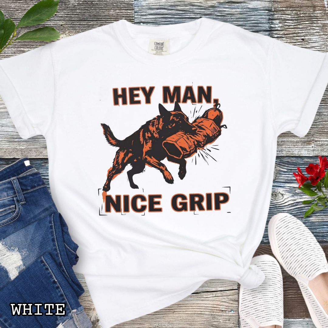 White Comfort Colors t-shirt with graphic design of a Schutzhund dog biting a sleeve and the humorous text 'Hey Man nice grip', shown on a wooden surface with accessories.