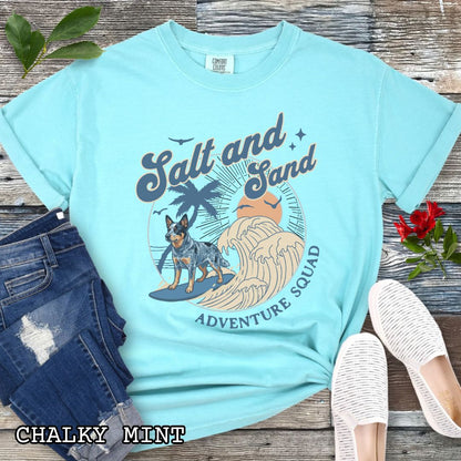 Mint Comfort Colors t-shirt for Australian Cattle Dog lovers with beach-themed graphic of a surfing Blue Heeler, waves and sunset, and the text 'Salt and Sand Adventure Squad' in retro lettering, shown on a wooden surface with accessories.