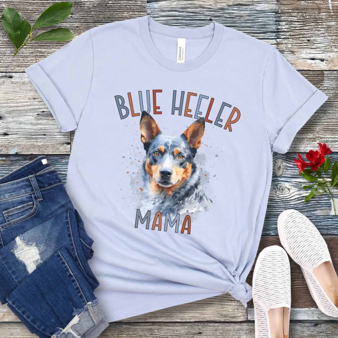 A light blue dog lover t-shirt with a watercolor style graphic of a Blue Heeler dog and the text 'Blue Heeler Mama' in colorful letters. The shirt is displayed accompanied by a pair of blue jeans and white shoes on a wooden surface.