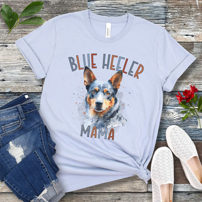 A light blue dog lover t-shirt with a watercolor style graphic of a Blue Heeler dog and the text 'Blue Heeler Mama' in colorful letters. The shirt is displayed accompanied by a pair of blue jeans and white shoes on a wooden surface.