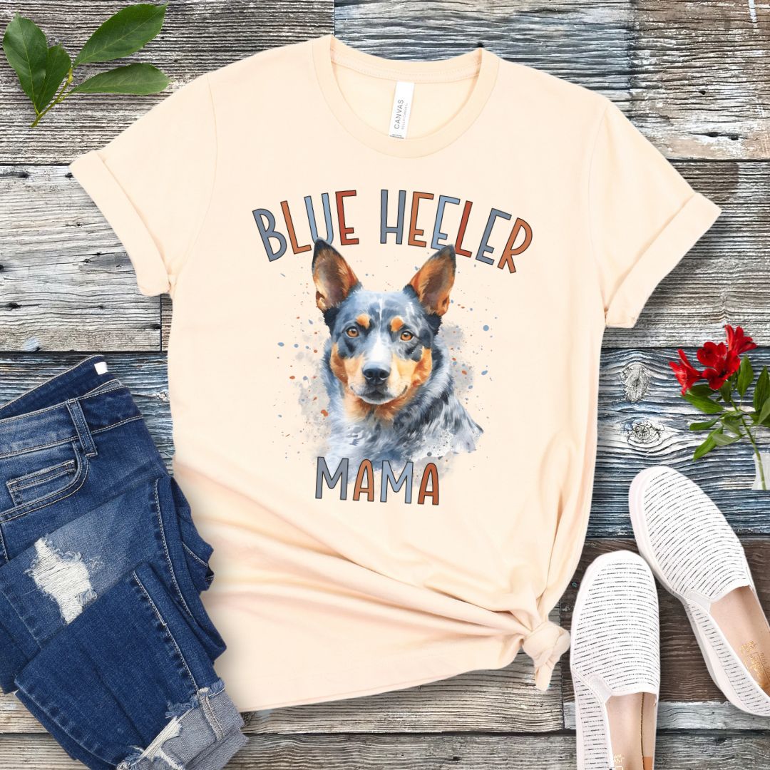 A beige dog lover t-shirt with a watercolor style graphic of a Blue Heeler dog and the text 'Blue Heeler Mama' in colorful letters. The shirt is displayed accompanied by a pair of blue jeans and white shoes on a wooden surface.