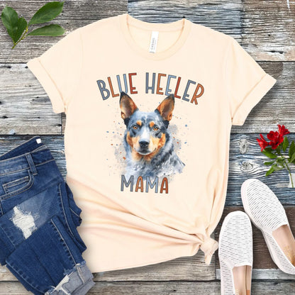 A beige dog lover t-shirt with a watercolor style graphic of a Blue Heeler dog and the text 'Blue Heeler Mama' in colorful letters. The shirt is displayed accompanied by a pair of blue jeans and white shoes on a wooden surface.