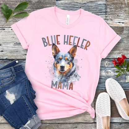 A pink dog lover t-shirt with a watercolor style graphic of a Blue Heeler dog and the text 'Blue Heeler Mama' in colorful letters. The shirt is displayed accompanied by a pair of blue jeans and white shoes on a wooden surface.