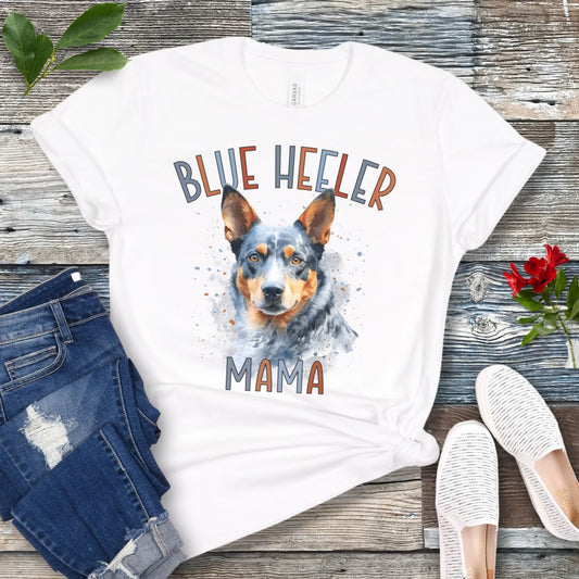 A white dog lover t-shirt with a watercolor style graphic of a Blue Heeler dog and the text 'Blue Heeler Mama' in colorful letters. The shirt is displayed accompanied by a pair of blue jeans and white shoes on a wooden surface.