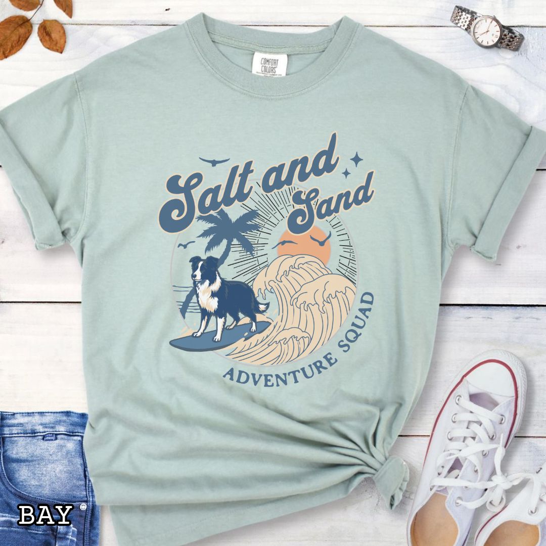Light green Comfort Colors t-shirt for Border Collie lovers with beach-themed graphic of a surfing GSD, waves and sunset, and the text 'Salt and Sand Adventure Squad' in retro lettering, shown on a wooden surface with accessories.