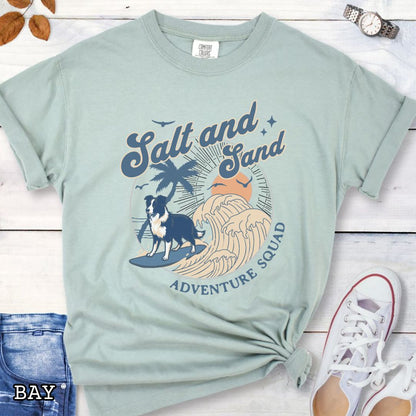 Light green Comfort Colors t-shirt for Border Collie lovers with beach-themed graphic of a surfing GSD, waves and sunset, and the text 'Salt and Sand Adventure Squad' in retro lettering, shown on a wooden surface with accessories.