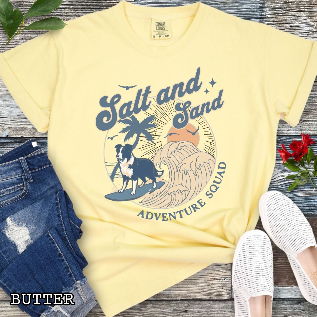 Yellow Comfort Colors t-shirt for Border Collie lovers with beach-themed graphic of a surfing GSD, waves and sunset, and the text 'Salt and Sand Adventure Squad' in retro lettering, shown on a wooden surface with accessories.