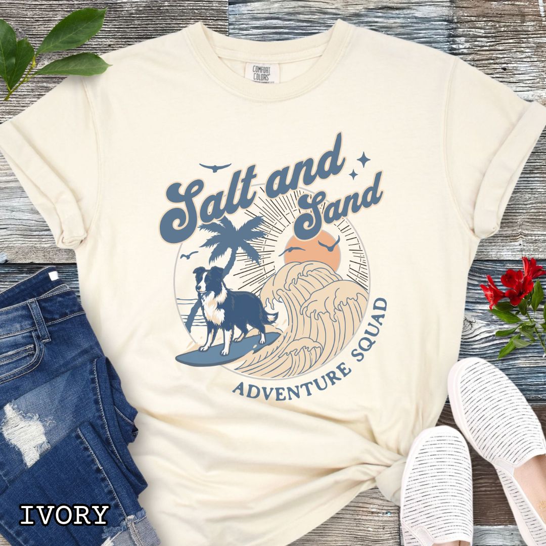 Beige Comfort Colors t-shirt for Border Collie lovers with beach-themed graphic of a surfing GSD, waves and sunset, and the text 'Salt and Sand Adventure Squad' in retro lettering, shown on a wooden surface with accessories.