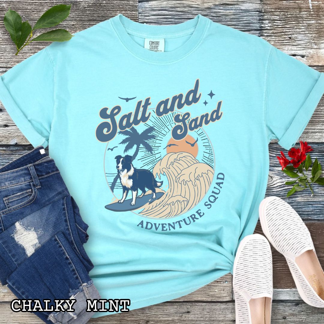 Mint Comfort Colors t-shirt for Border Collie lovers with beach-themed graphic of a surfing GSD, waves and sunset, and the text 'Salt and Sand Adventure Squad' in retro lettering, shown on a wooden surface with accessories.
