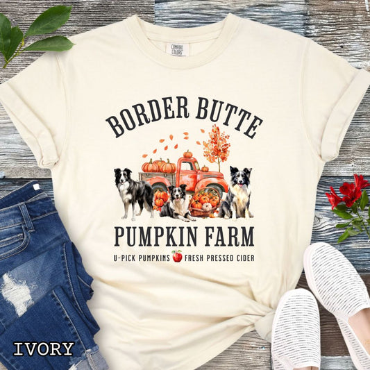 Beige fall-theme t-shirt for Border Collie lovers with 'Border Butte Pumpkin Farm' design featuring Border Collies, fall foliage and a pumpkin truck, shown on a wooden surface.