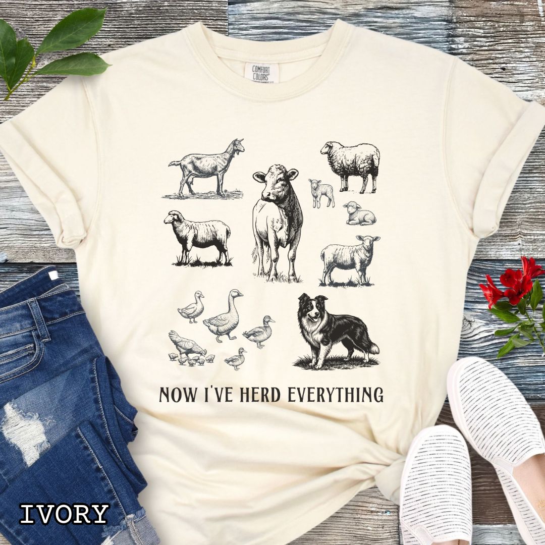 Beige Comfort Colors t-shirt for Border Collie lovers with vintage farm animal illustrations and a Border Collie dog, with the humorous text 'Now I've Herd Everything', shown on a wooden surface with denim jeans and flowers.