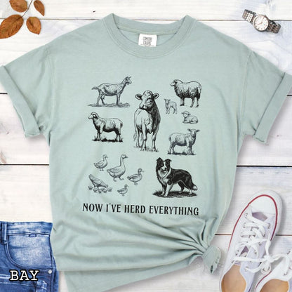 Light green Comfort Colors t-shirt for Border Collie lovers with vintage farm animal illustrations and a Border Collie dog, with the humorous text 'Now I've Herd Everything', shown on a wooden surface with denim jeans and flowers.