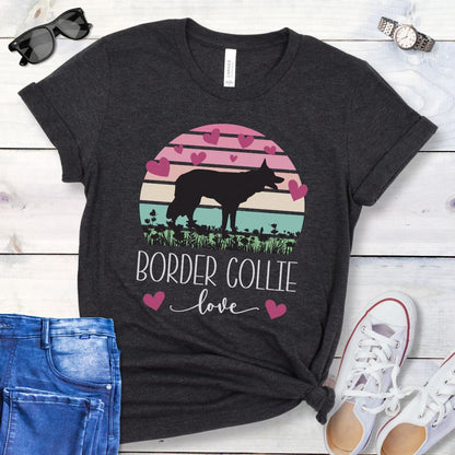 Dark grey t-shirt with Border Collie silhouette amid pastel vintage sunset and hearts, with the text 'Border Collie love'. Shown on a wooden surface with denim shorts and white shoes.
