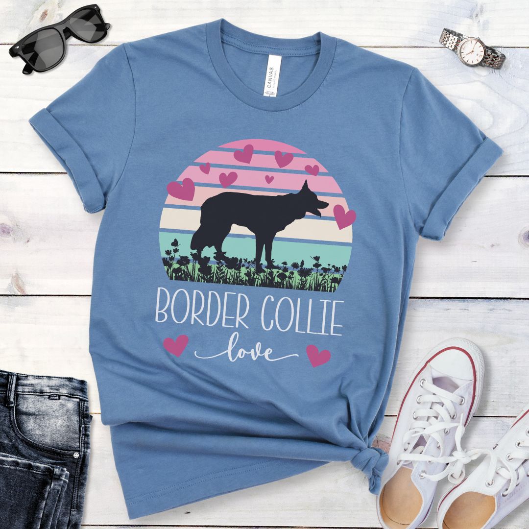 Blue t-shirt with Border Collie silhouette amid pastel vintage sunset and hearts, with the text 'Border Collie love'. Shown on a wooden surface with denim shorts and white shoes.
