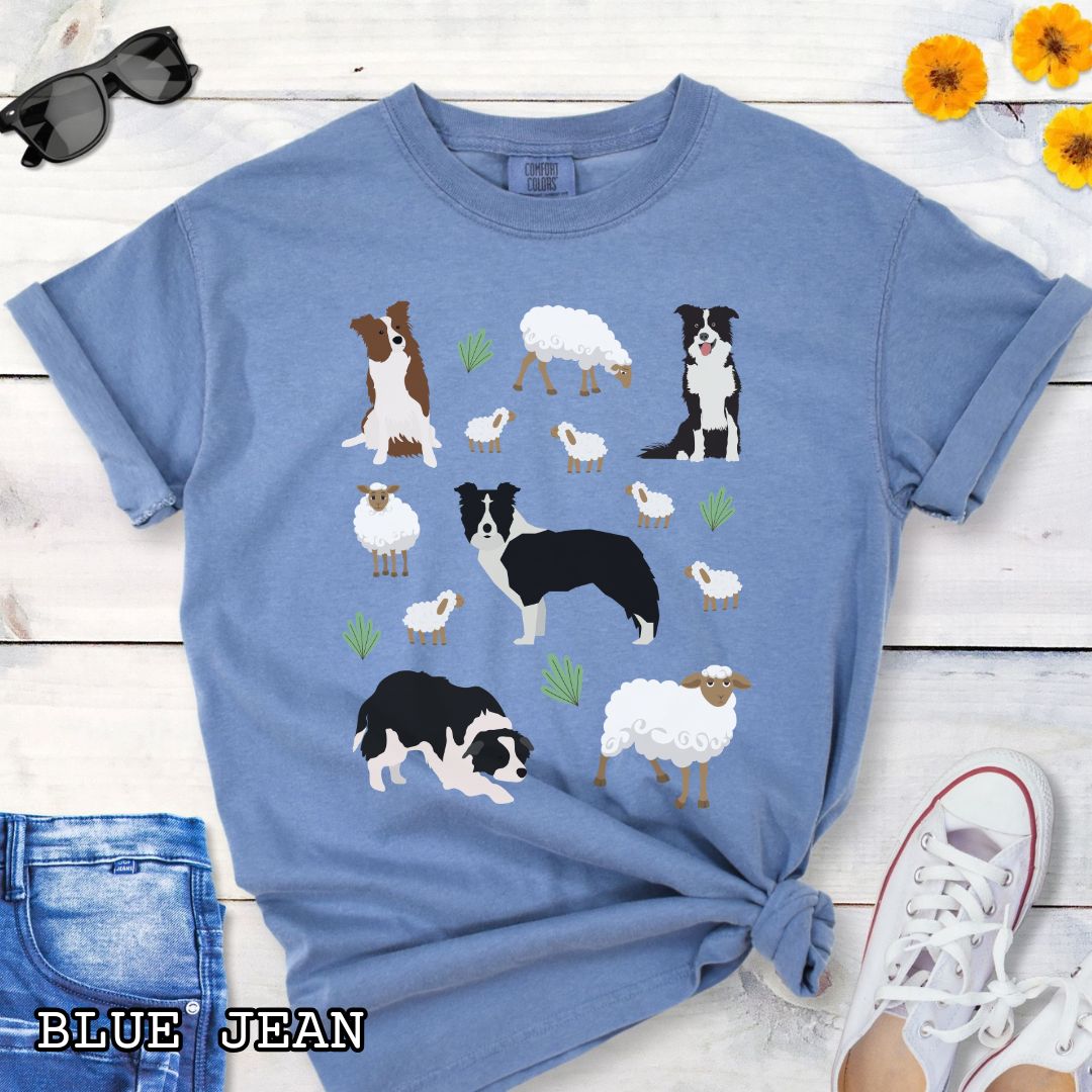 Blue Comfort Colors  t-shirt with Border Collie dogs and grazing sheep, done in Cottage Core style, shown on a wooden surface with sunglasses and flowers.