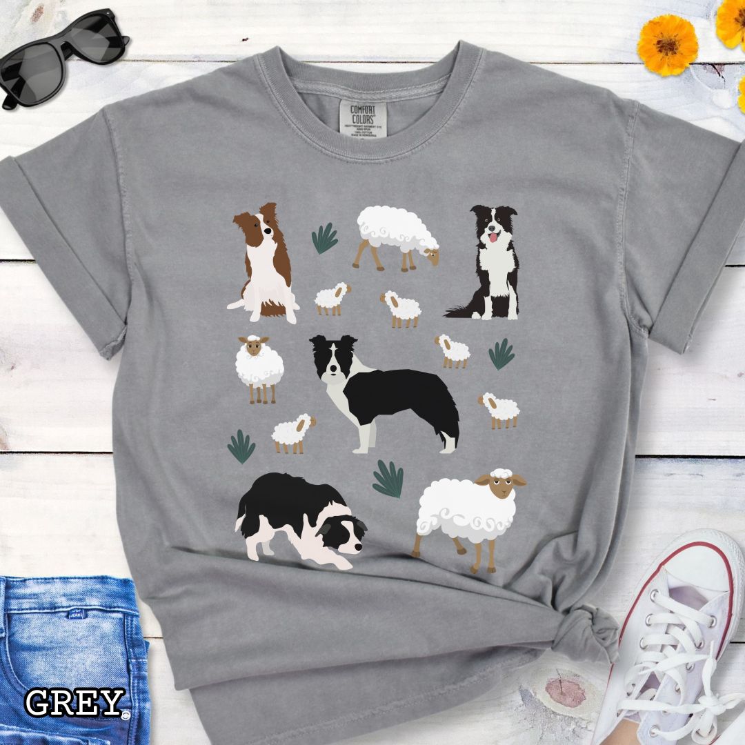 Grey Comfort Colors  t-shirt with Border Collie dogs and grazing sheep, done in Cottage Core style, shown on a wooden surface with sunglasses and flowers.