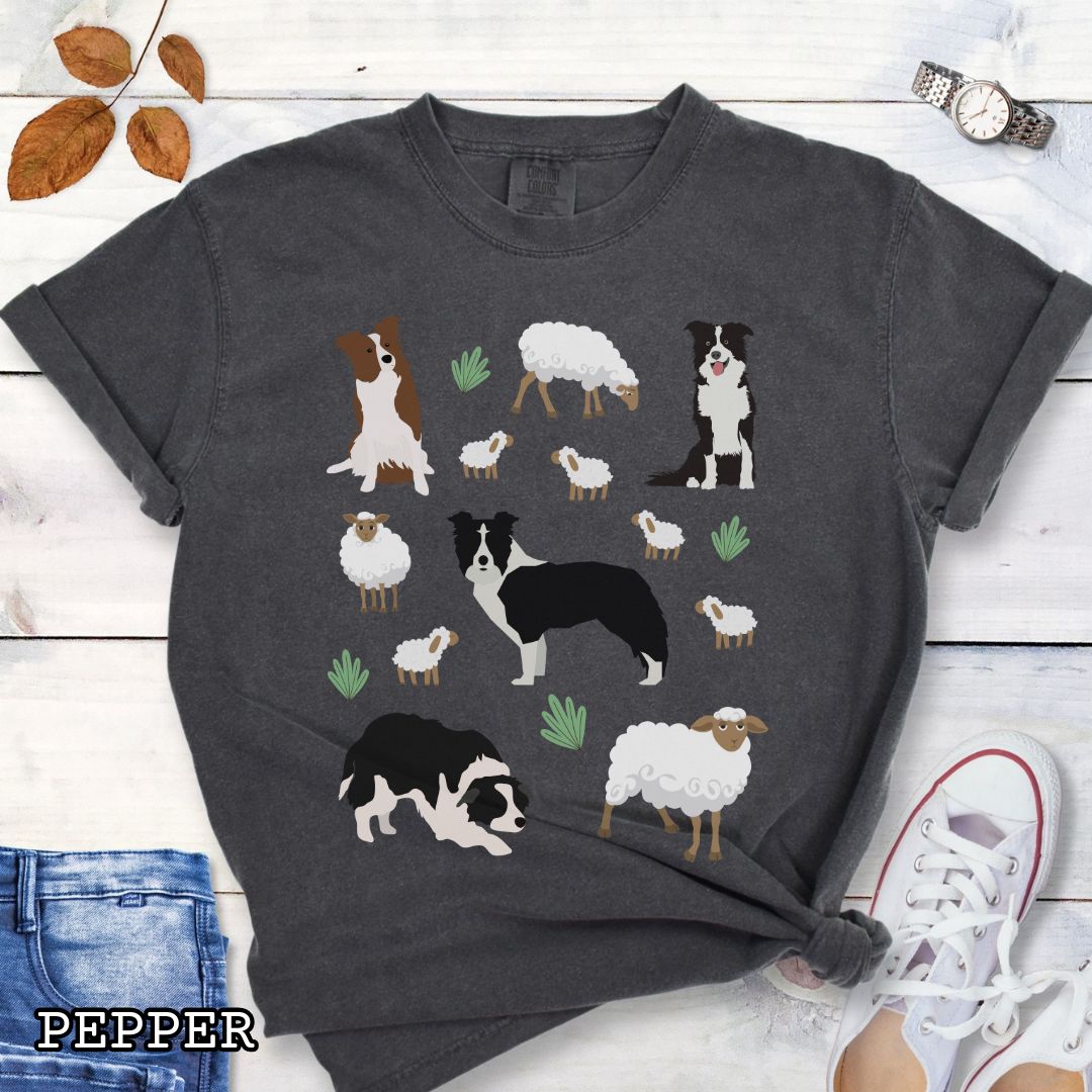 Dark grey Comfort Colors  t-shirt with Border Collie dogs and grazing sheep, done in Cottage Core style, shown on a wooden surface with sunglasses and flowers.