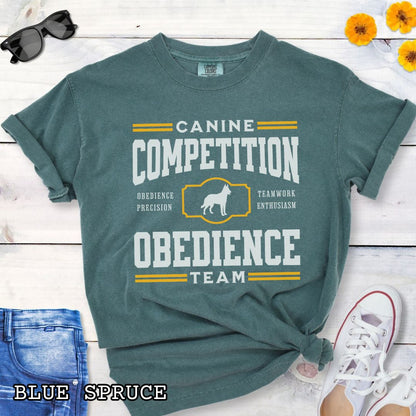 Green Comfort Colors dog obedience t-shirt with 'Canine Competition Obedience Team' text and custom breed silhouette, presented on a wooden surface with denim jeans and a flower.