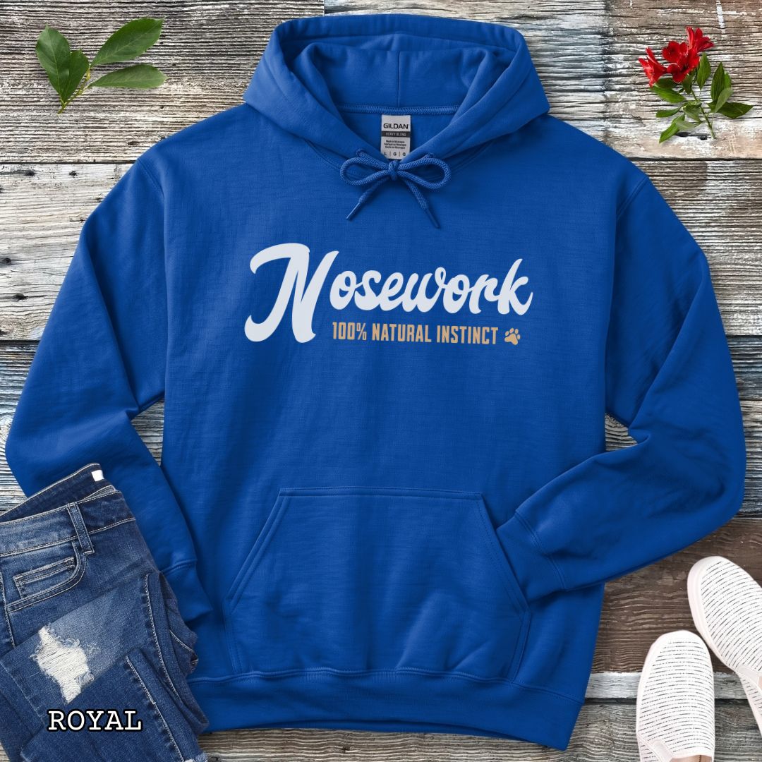 A royal blue pullover hoodie for canine scent detection trainers, laid flat on a rustic wood surface, with white and tan text reading 'Nosework 100% Natural Instinct' across the chest. The hoodie is styled with blue jeans, white shoes, green leaves, and red flowers around it.