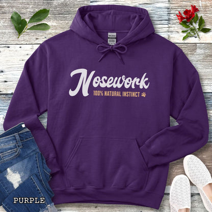 A purple pullover hoodie for canine scent trainers, laid flat on a rustic wood surface, with white and tan text reading 'Nosework 100% Natural Instinct' across the chest. The hoodie is styled with blue jeans, white shoes, green leaves, and red flowers around it.