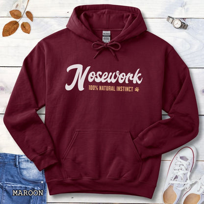 A maroon pullover hoodie for canine scent trainers, laid flat on a rustic wood surface, with white and tan text reading 'Nosework 100% Natural Instinct' across the chest. The hoodie is styled with blue jeans, white shoes, green leaves, and red flowers around it.