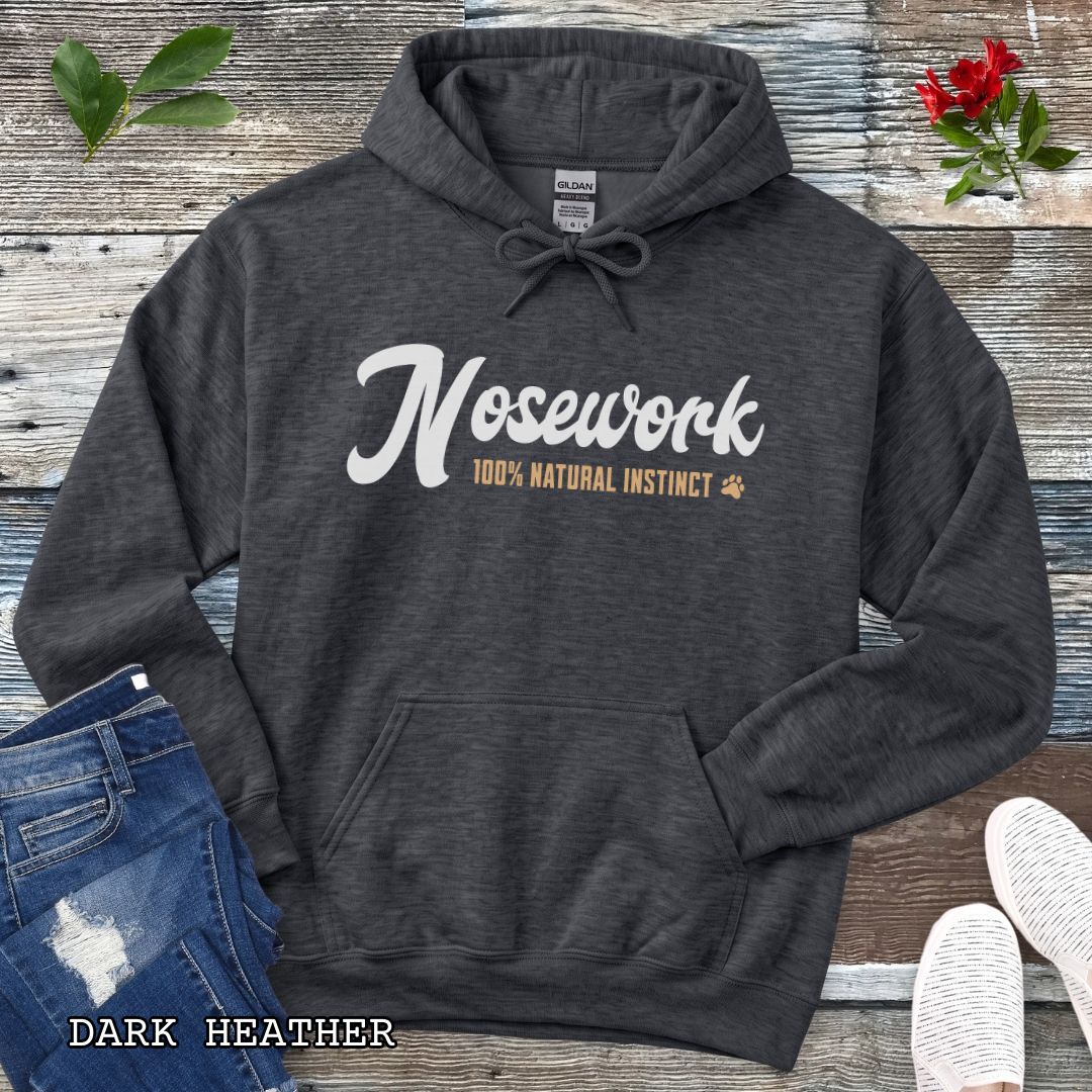 A dark grey pullover hoodie for canine scent trainers, laid flat on a rustic wood surface, with white and tan text reading 'Nosework 100% Natural Instinct' across the chest. The hoodie is styled with blue jeans, white shoes, green leaves, and red flowers around it.