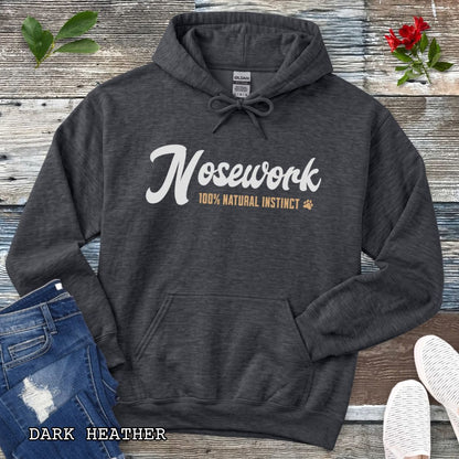 A dark grey pullover hoodie for canine scent trainers, laid flat on a rustic wood surface, with white and tan text reading 'Nosework 100% Natural Instinct' across the chest. The hoodie is styled with blue jeans, white shoes, green leaves, and red flowers around it.