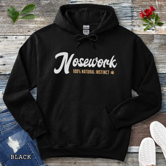 A black pullover hoodie for canine scent trainers, laid flat on a rustic wood surface, with white and tan text reading 'Nosework 100% Natural Instinct' across the chest. The hoodie is styled with blue jeans, white shoes, green leaves, and red flowers around it.