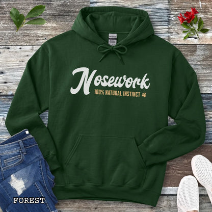 A dark green pullover hoodie for canine scent trainers, laid flat on a rustic wood surface, with white and tan text reading 'Nosework 100% Natural Instinct' across the chest. The hoodie is styled with blue jeans, white shoes, green leaves, and red flowers around it.