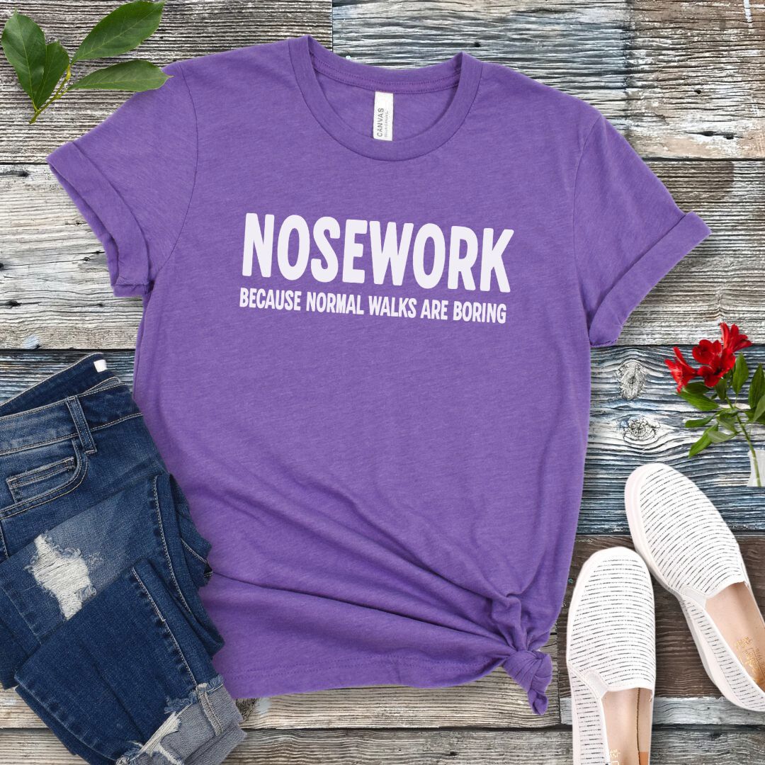 Navy blue canine scent training t-shirt with 'NOSEWORK BECAUSE NORMAL WALKS ARE BORING' text on a wooden surface with jeans and shoes.