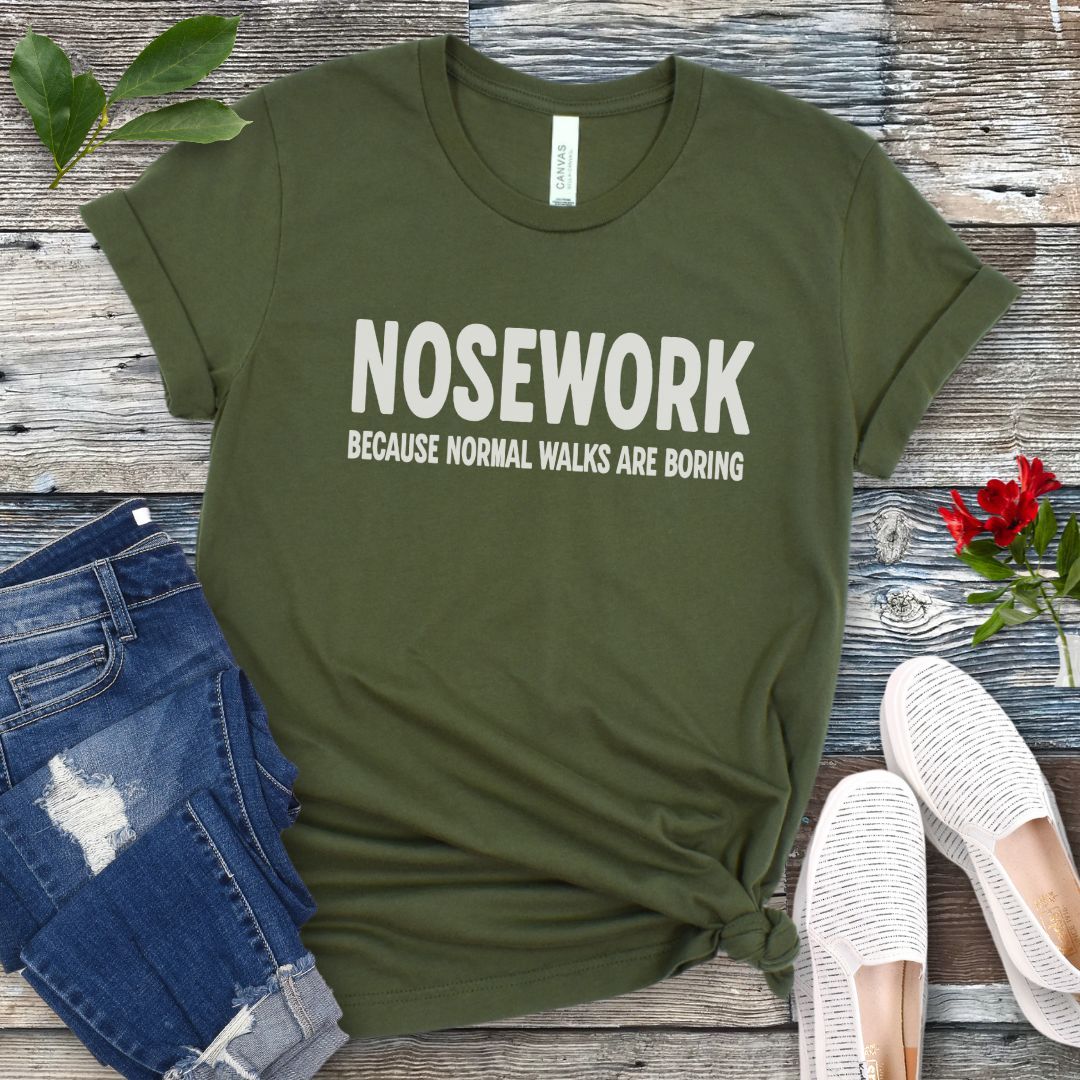 Military green K9 Nose Work t-shirt with 'NOSEWORK BECAUSE NORMAL WALKS ARE BORING' text on a wooden surface with jeans and shoes.